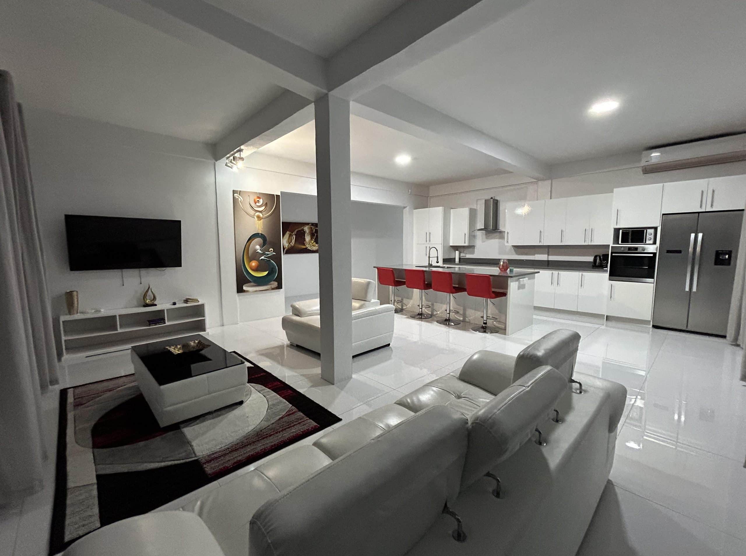Modern open-plan living area with white furniture, kitchen island with red bar stools, abstract wall art, flat-screen TV, and sleek white cabinetry. The room is brightly lit with a minimalist design.